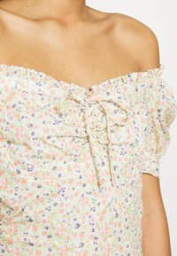 Kobieta w białej sukience odkrywającej ramiona, ozdobionej małymi różowymi, fioletowymi i zielonymi wzorami kwiatowymi oraz związaną kokardą na piersi.