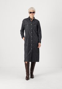Džínová košeľová šaty v tmavom práve s prednými gombíkmi, dvomi náprsními vreckami, dlhými rukávmi a bočnými vreckami, doplnené hnedými čižmami po kolená.