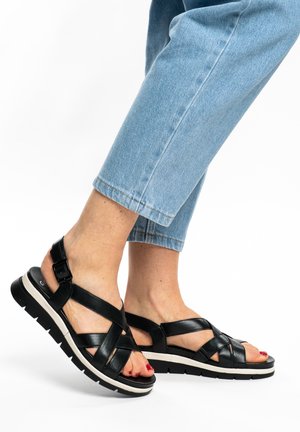 Pies con sandalias negras de tiras y suelas gruesas, combinadas con jeans cortos de mezclilla azul claro sobre un fondo blanco.