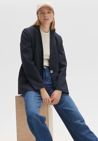 Blazer bleu foncé avec un bouton unique, associé à un top beige clair et un jean bleu à jambe large. Le mannequin porte une casquette beige. Texture de tissu lisse.