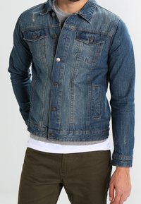 Blå denimjacka med klassisk skärning, med två framfickor, metallknappar och subtila slitna detaljer. Bärs över en grå tröja.