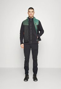 Moška pohodniška obleka z črno flis jakno z zadrgo, brezrokavnikom z zelenimi poudarki, črnimi hlačami in robustnimi črnimi čevlji.