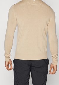 Man som bär en beige långärmad tröja och svarta byxor, sett bakifrån med avslappnade händer vid sidorna mot en neutral bakgrund.