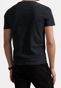 Man som bär en mörkgrå t-shirt med kort ärm och svarta byxor, sedd från ryggen mot en enkel ljus bakgrund.