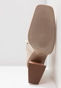 Chaussure à talon beige pour femme avec bout carré et semelle texturée, positionnée à la verticale sur une surface blanche contre un fond clair.