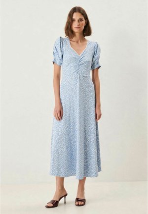 Nainen seisoo päällään vaaleansininen, kukallinen midimekko, jossa on rypytetyt lyhyet hihat, ja ruskeat avonaiset nilkkurit kannalla yksiväristä taustaa vasten.