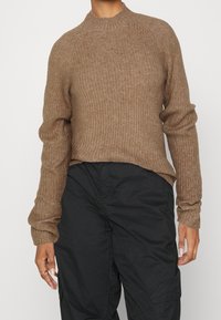 Brown ribbed sweater with a high neckline and long sleeves, featuring a relaxed fit and textured fabric, paired with black pants.