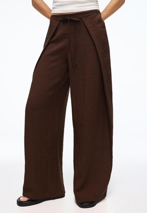 Kvinde iført højtaljede, viddebrune bukser med bindebånd foran og lommer, kombineret med sorte sandaler og en hvid top.