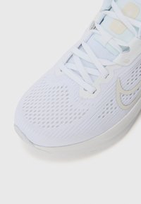 Bílé sportovní boty s prodyšným síťovaným svrškem, texturovaným designem, světle modrými akcenty a flexibilní podrážkou. Obsahují bílé tkaničky a branding.