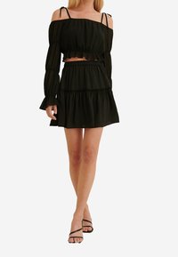 Crop top nero off-the-shoulder con maniche lunghe e dettagli a volant, abbinato a una mini gonna a volant coordinata. Entrambi in un tessuto trasparente.