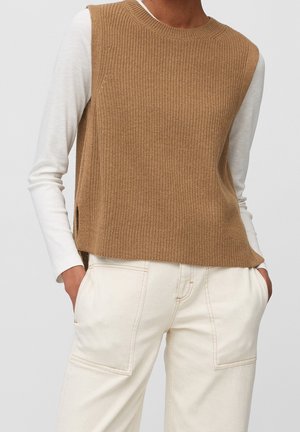 Persona que lleva un chaleco de punto sin mangas color marrón camello sobre una camiseta blanca de manga larga, combinada con pantalones de talle alto en color blanco roto con grandes bolsillos delanteros.