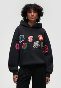 Hoodie nero con lettere ricamate colorate in stili diversi. Il design include una tasca frontale e polsini a costine. Tessuto morbido.
