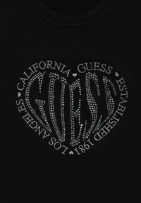 Svart topp med hjerteformet design pyntet med sølvstrass. Tekst inkluderer "Guess," "California," og "Etablert 1981."