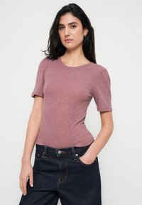 Πλεκτό εφαρμοστό τοπ με ραβδώσεις σε χρώμα σκουριασμένο ροζ, με κοντά puff μανίκια, στρογγυλή λαιμόκοψη και κυματιστή λεπτομέρεια στο τελείωμα, συνδυασμένο με σκουρόχρωμο τζιν παντελόνι.