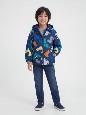 Smilende dreng iført en blå hættetrøje med dinosaurprint, grå t-shirt, blå jeans og marineblå slip-on sko, stående med den ene hånd i lommen.