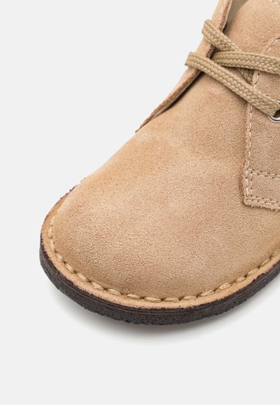 Pantof suede bej cu suprafață texturată, vârf rotund și șireturi bej. Dispune de cusături tonale în jurul tălpii din cauciuc.
