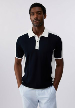 Uomo che indossa una polo a maniche corte bianca e nera con bottoni bianchi, in piedi con le mani nelle tasche davanti a uno sfondo semplice.