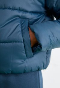 Modrá pu puffová bunda s hladkým, lesklým povrchom; má bočnú kapsu a rebrované manžety; názov značky "THERM" je viditeľný na rukáve.