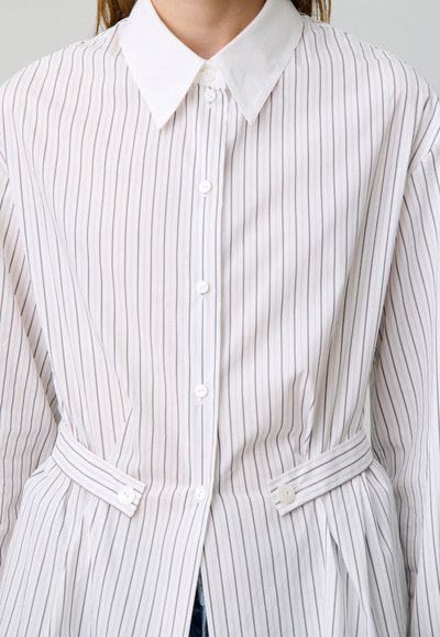 Camicia a righe bianche con colletto classico, chiusura a bottoni e vita con cintura e bottoni decorativi. Tessuto morbido con linee verticali.