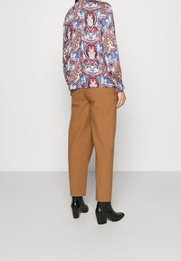 Blusa a maniche lunghe con un motivo paisley colorato, abbinata a pantaloni marroni a sigaretta e stivaletti neri con tacco. Sfondo neutro.