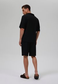 Črna polo majica s kratkimi rokavi, v kombinaciji s črnimi kratkimi hlačami. Model je viden s hrbta, nosi rjave natikače. Gladek, minimalističen dizajn.