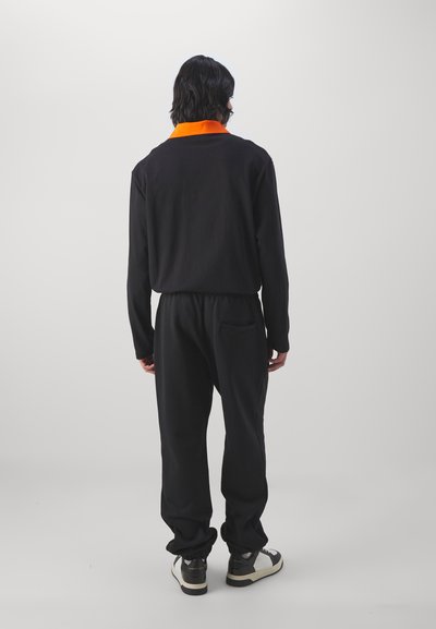 Sort langærmet bluse med en orange krave, parret med sorte joggerbukser. Blød tekstur, afslappet pasform, set fra ryggen, båret sammen med sort og hvidt sneakers.