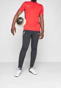 Muž v červeném sportovním tričku a černých kalhotách Liverpool FC drží černý a zlatý fotbal na boku, stojí na jednobarevném pozadí.