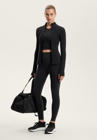 Kobieta stoi, ubrana w czarną sportową kurtkę, krótki top, legginsy i trampki, trzymając dużą czarną torbę gymnastyczną na tle jednolitym.