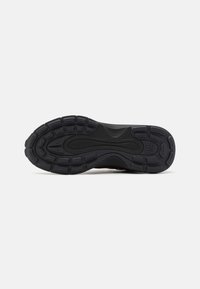 Black athletic shoe sole with a textured rubber surface, featuring a wavy pattern and raised circular grips for traction.