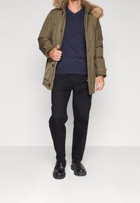 Oliven grøn parka med pelsforet hætte, parret med en marineblå V-hals skjorte og sorte bukser. Sorte ankelstøvler med en robust sål.