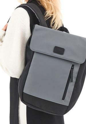 Une personne blonde portant une tenue noire et blanche, portant un sac à dos rectangulaire gris et noir avec une poche à fermeture éclair à l'avant.