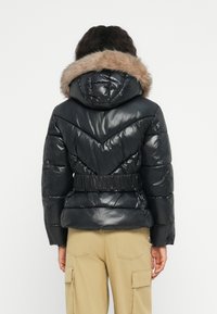 Person in einer schwarzen glänzenden Pufferjacke mit fellgefütterter Kapuze und elastischem Gürtel, kombiniert mit beigen Cargo-Hosen, die mit dem Rücken zur Kamera steht.
