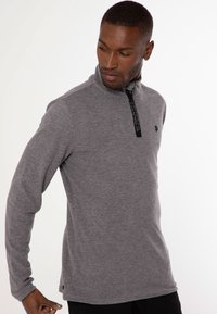 Grå langærmet pullover med lynlås i kraven, udstyret med et sort logo på brystet og tekstureret stof for komfort.
