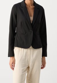 Schwarzer taillierter Blazer mit einem Knopf, Reverskragen und zwei vorderen Taschen, kombiniert mit hellen, strukturierten, crema-farbenen Hosen.