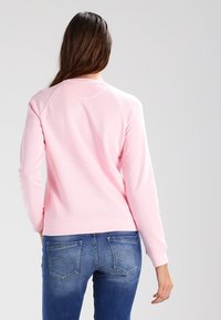 Felpa rosa a maniche lunghe, con polsini e orlo a costine. Presenta un colletto rotondo e un tessuto morbido e testurizzato. Abbinata a jeans blu.