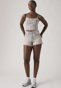 Γυναίκα που στέκεται με τα χέρια πίσω από την πλάτη, φορώντας ανοιχτόχρωμο κολλώδες crop top από τζιν, ταιριαστά κοντά σκισμένα σορτς, λευκές κάλτσες και ασημένια αθλητικά παπούτσια.