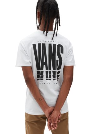 Persoon met gevlochten haar, gekleed in een witte Vans-t-shirt met een groot zwart logo en tan kleurige broek, staande met de handen samengevouwen achter de rug.