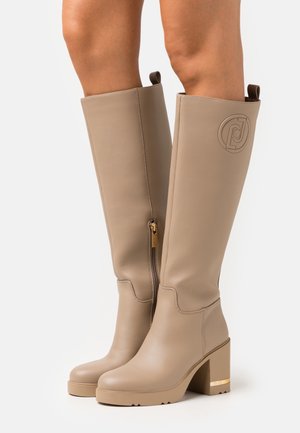Beige nahkäbootsit, jotka ulottuvat polven ylle. Niissä on leveä korko, sivuvetoketjut, vetotapit ja kohokuvioitu ympyrälogo ulkopuolella. Ne on yhdistetty paljaisiin jalkoihin.