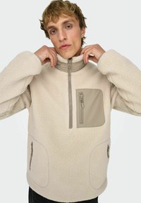 Beige, pehmeä fleece-pulloveri, jossa on korkea kaulus, puolivetoketjullinen etuosa ja vetoketjullinen tasku. Sivutaskut vetoketjuilla.