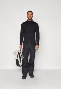 Veste noire zippée, pantalon noir et chaussures noires. Tient un sac à dos beige et noir. Texture lisse, design ajusté, détails minimalistes.