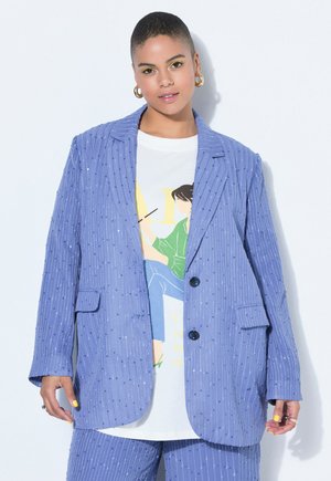 Jeune femme portant un blazer et un pantalon à fines rayures violettes claires, un t-shirt blanc à motif, des boucles d'oreilles en or en forme de créole et un maquillage des yeux jaune vif.