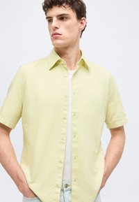 Jeune homme aux cheveux courts et bouclés portant une chemise à manches courtes jaune clair boutonnée par-dessus un t-shirt blanc et un jean bleu clair, les mains dans les poches.
