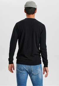 Man med grå stickad mössa, svart långärmad tröja och blå jeans, står vänd bort från kameran mot en enkel ljus bakgrund.