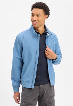 Jonge man die glimlacht, draagt een lichtblauw jack met rits over een donker poloshirt en grijze cargobroek, staand tegen een witte achtergrond.