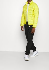 Yellow padded jacket with a stand-up collar, zip closure, and ribbed cuffs, paired with black sweatpants and white sneakers.