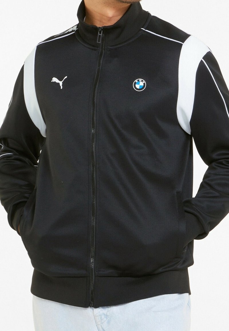 Veste zippée noire avec des accents latéraux blancs, arborant un logo Puma et un écusson BMW sur la poitrine. Fabriquée dans un matériau lisse et léger.