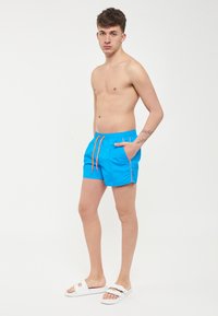 Pantaloni da mare blu con cintura elastica e cordoncino arancione, tasca laterale e cuciture a contrasto; tessuto liscio e leggero.