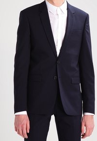 Mörkblå kostym med en skräddarsydd passform, som har en enfärgad kavaj, två framfickor och slagkrage, kombinerad med en vit skjorta.