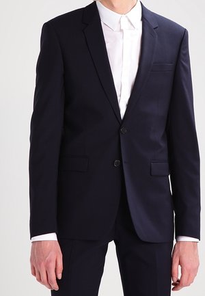 Costume bleu marine avec une coupe ajustée, comprenant une veste à une seule boutonnière, deux poches à l'avant et un revers cranté, associé à une chemise blanche.