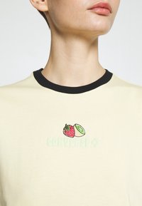 Kremowa koszulka z czarnym okrągłym dekoltem. Ma na środku grafikę truskawki i kiwi, a poniżej napis "CONVERSE" w jasnej zieleni.